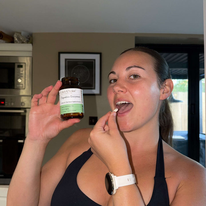 A woman in a kitchen setting holds up an amber supplement bottle labeled 'Udo's Choice Digestive Enzyme' while taking a white tablet from her finger. She is wearing a black halter top and a white smartwatch.