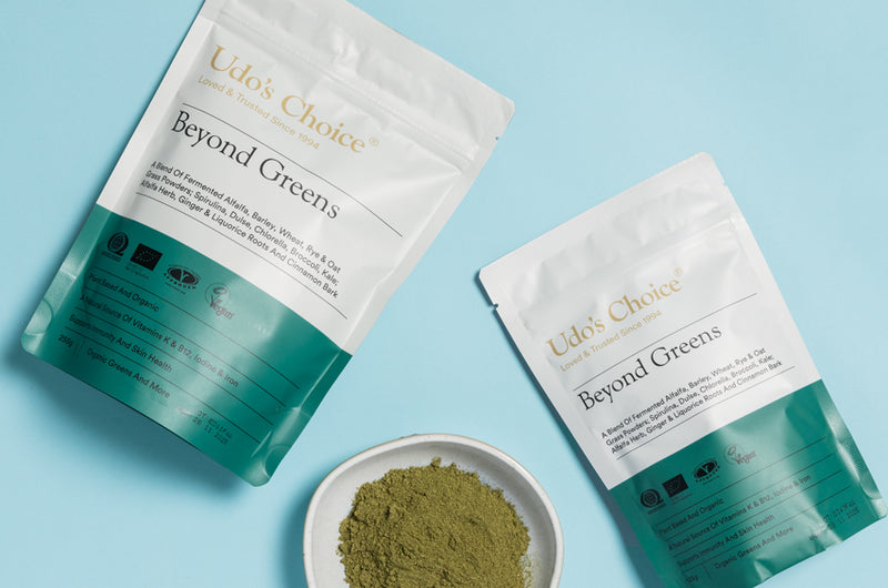 Two packages of 'Udo's Choice Beyond Greens supergreens powder' on a light blue background with a small bowl of green powder.