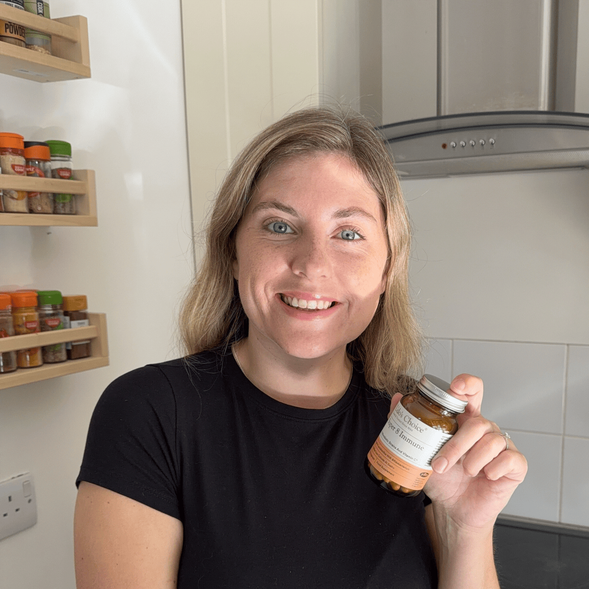 A smiling woman with blonde hair wearing a black t-shirt holds up a bottle of Super Immune 8 Probiotics with Vitamin C in a kitchen setting.