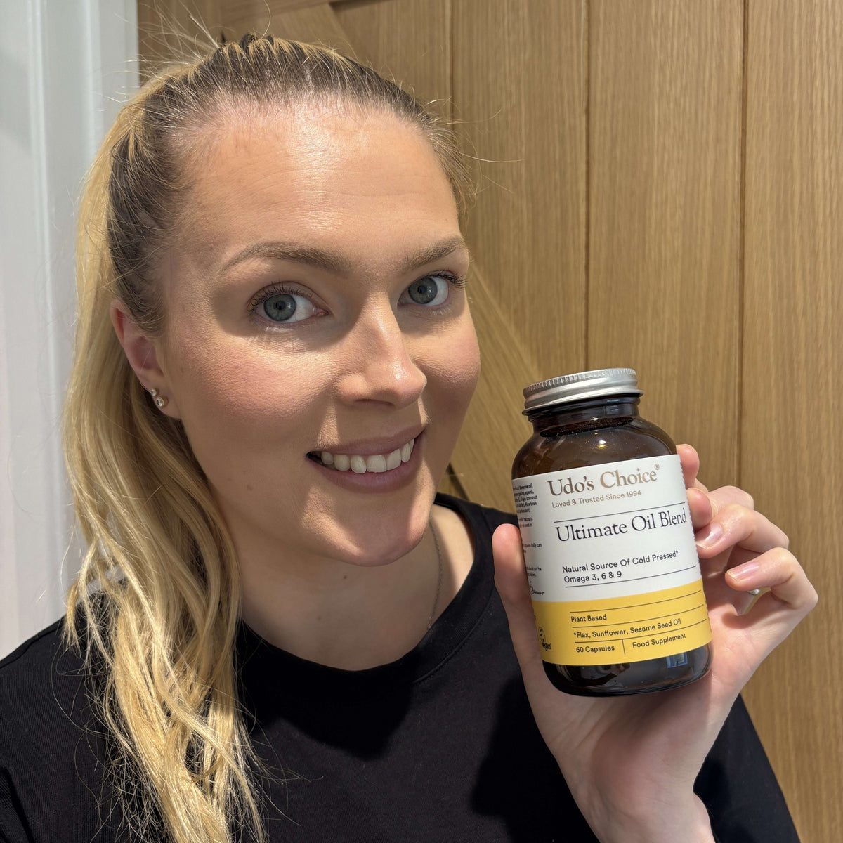 A smiling blonde woman holds a bottle of Udo's Choice Ultimate Oil Blend in her hand, set against a background of light wood paneling.