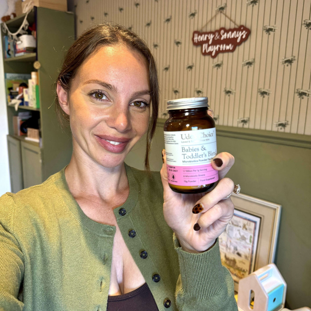 A woman holding a bottle of Udo's Choice Babies & Toddlers Probiotic supplement in front of children's nursery wall.