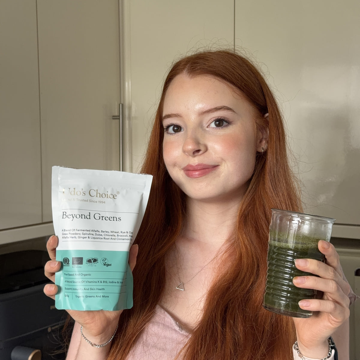 A red haired girl holding a pack of Udo's Choice Beyond Greens supergreens powder and a glass of the supergreens vegan powder for immunity and skin mixed into water