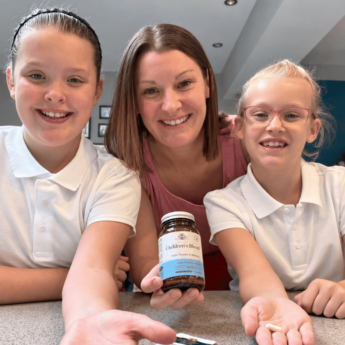 A woman and two smiling girls are gathered around a table, with the woman holding a bottle labeled Children's Blend Probiotic supplement. The girls are wearing white polo shirts, and the setting appears to be an indoor home environment.