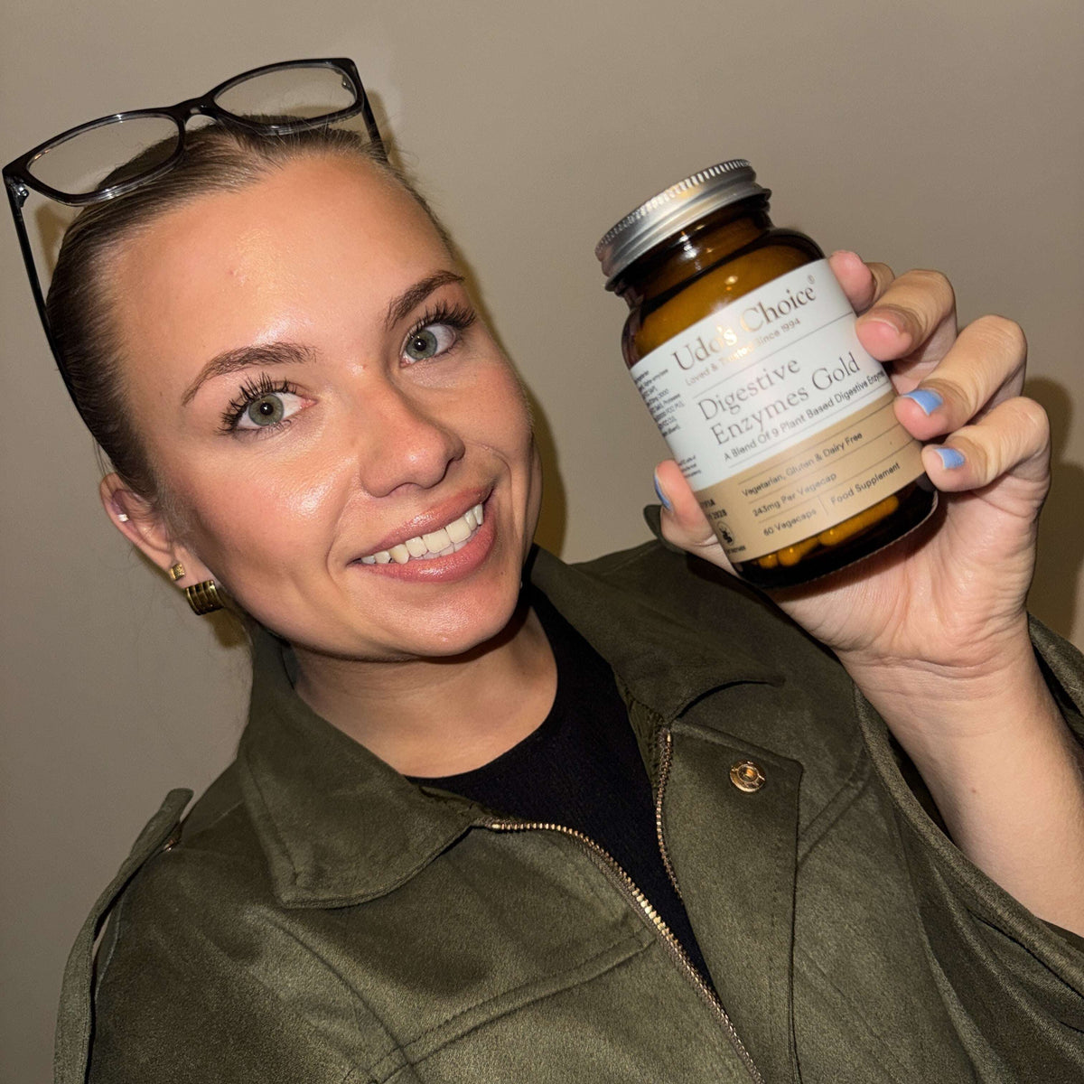 A woman holding a bottle of Udo's Choice Digestive Enzymes Gold supplement, wearing an olive green jacket and glasses on her head against a plain beige wall.